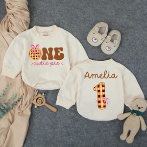 May include: Two cream-colored baby bodysuits with long sleeves. One says "ONE cutie pie" with a pie graphic. The other says "Amelia" and has a number one graphic. Also shown are bear slippers and a teddy bear.