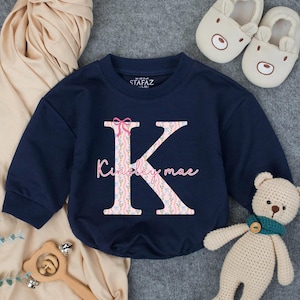 May include: Navy blue baby sweatshirt with a large floral-patterned letter "K" and the name "Kinley mae" in pink script. The sweatshirt has a pink bow at the top of the letter. The brand name "STAFZ" is printed on the collar.