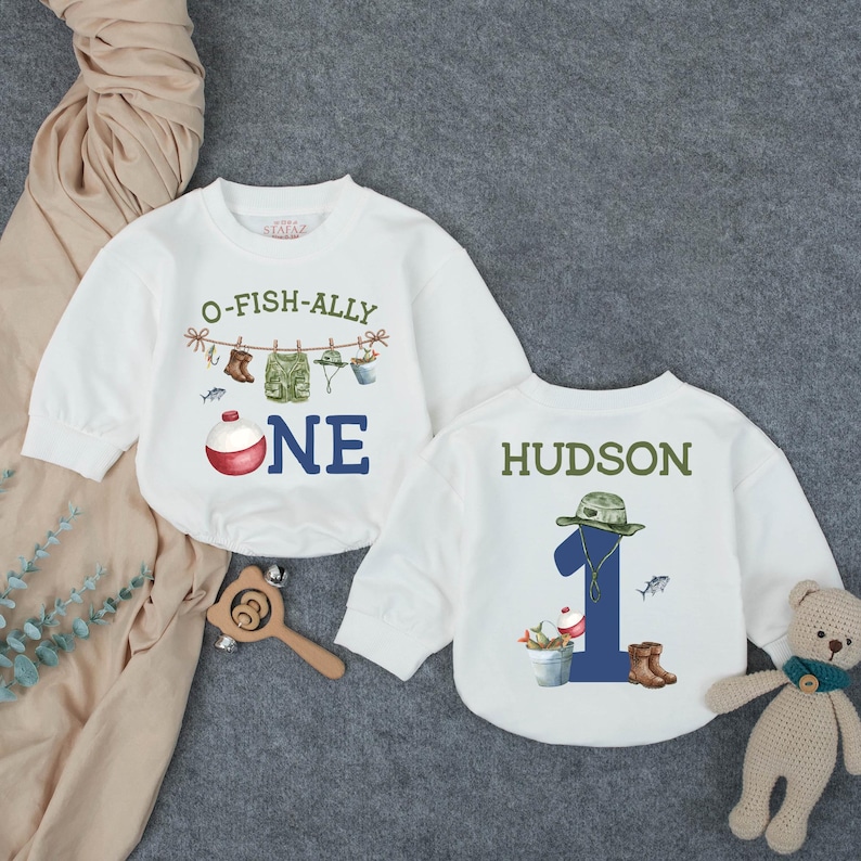 May include: Two white long-sleeve shirts featuring fishing-themed graphics. One shirt displays the text O-FISH-ALLY ONE with a fishing line, boots, vest, hat, and bobber. The other shirt shows the name HUDSON above a large blue number 1.