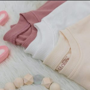 Puede incluir: Tres sudaderas de colores pastel: rosa empolvado, blanco y melocot&oacute;n, est&aacute;n cuidadosamente dobladas. Dos malvaviscos en forma de coraz&oacute;n y un collar de cuentas de madera est&aacute;n dispuestos sobre una superficie blanca y esponjosa. La sudadera melocot&oacute;n tiene una peque&ntilde;a etiqueta.