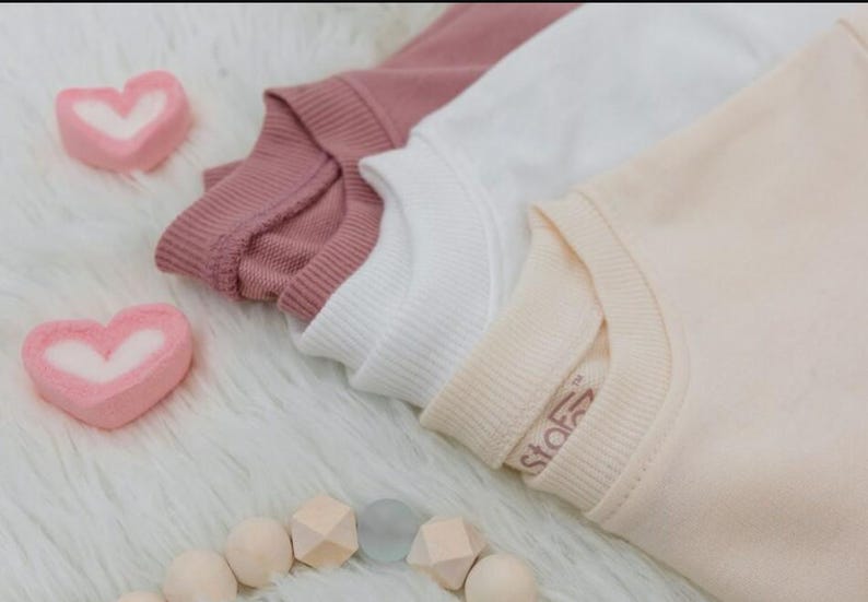 Puede incluir: Tres sudaderas de colores pastel: rosa empolvado, blanco y melocot&oacute;n, est&aacute;n cuidadosamente dobladas. Dos malvaviscos en forma de coraz&oacute;n y un collar de cuentas de madera est&aacute;n dispuestos sobre una superficie blanca y esponjosa. La sudadera melocot&oacute;n tiene una peque&ntilde;a etiqueta.