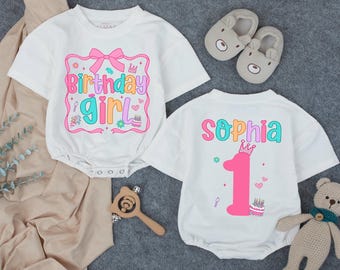 May include: Two white baby rompers with colorful graphics. One says "Birthday Girl" with a pink bow and the other says "Sophia" with a number 1 and a crown. Accessories include bear slippers and a stuffed bear.