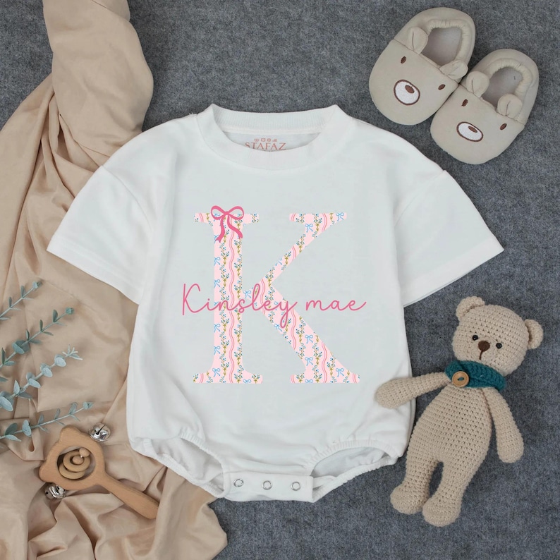 May include: White baby onesie with a large floral "K" initial and the name "Kinsley mae" in pink script. A pink bow is at the top of the letter. The image includes a pair of bear-shaped baby slippers and a small teddy bear.