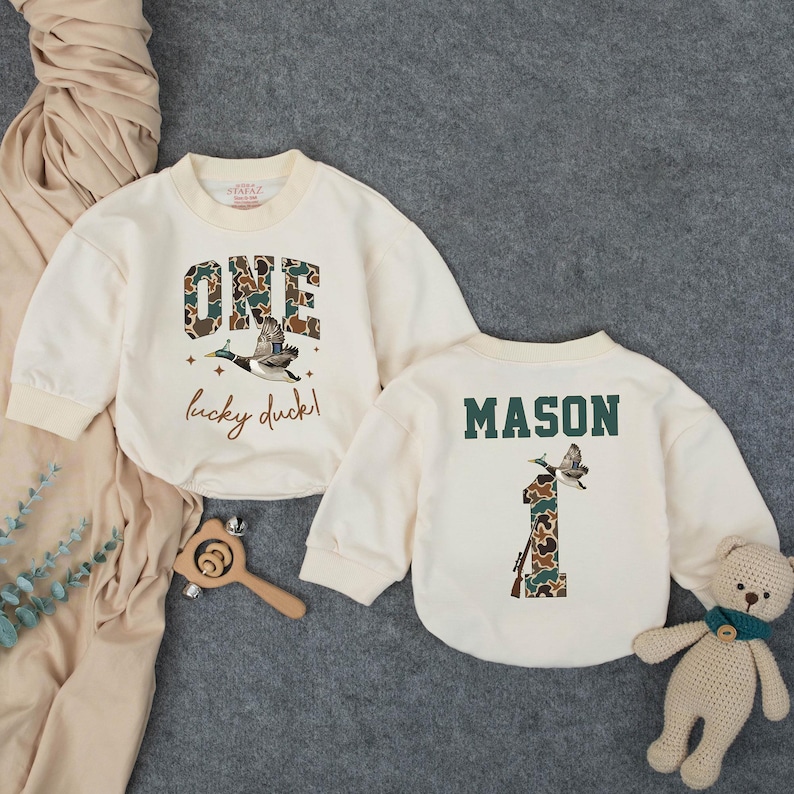 Puede incluir: Dos sudaderas crema para beb&eacute;s. Una presenta la palabra "ONE" en estampado de camuflaje con un gr&aacute;fico de pato volador y el texto "lucky duck!". La otra tiene el nombre "MASON" y el n&uacute;mero "1" en estampado de camuflaje con un gr&aacute;fico de pato.