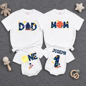 May include: Two white short-sleeve t-shirts and two white short-sleeve bodysuits featuring space-themed graphics. The shirts display the words DAD and MOM with astronaut and planet designs. The bodysuits feature the words ONE and JOSEPH with a number 1.