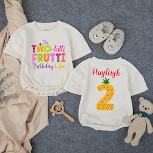 May include: Two cream-colored baby rompers with colorful text and graphics. One says "The Two-tutti Frutti Birthday Cutie." The other has a pineapple graphic with the number "2" and the name "Hayleigh." Also pictured are baby shoes, a wooden rattle, and a teddy bear.