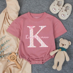 May include: A dusty rose baby romper with a large floral-patterned letter "K" and the name "Kinsley mae" printed on the front. The romper has short sleeves and snap closures. A small crocheted teddy bear and bear-shaped baby slippers are next to the romper.