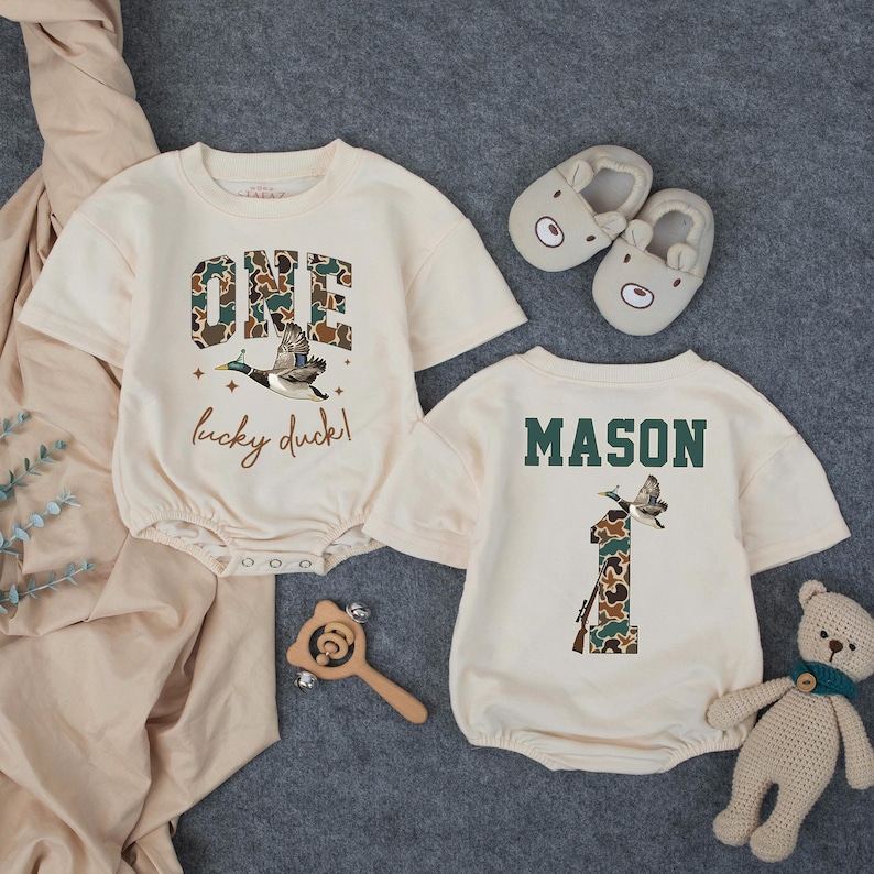 Puede incluir: Dos peleles de beb&eacute; color crema con mangas cortas. Uno dice "ONE lucky duck!" con un gr&aacute;fico de pato y letras de camuflaje. El otro dice "MASON" y tiene un gr&aacute;fico de pato y el n&uacute;mero "1" en camuflaje. Tambi&eacute;n hay un par de zapatillas de oso y un osito de peluche.