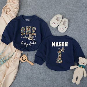 Puede incluir: Dos sudaderas azul marino con el texto "ONE lucky duck!" y "MASON". El n&uacute;mero "1" tambi&eacute;n aparece en la parte posterior de una sudadera. Las sudaderas tienen un dise&ntilde;o de pato y camuflaje. Un par de zapatos de beb&eacute; blancos y un oso de peluche tambi&eacute;n est&aacute;n en la imagen.
