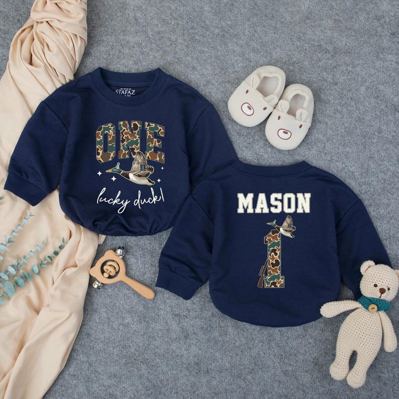 Puede incluir: Dos sudaderas azul marino con el texto "ONE lucky duck!" y "MASON". El n&uacute;mero "1" tambi&eacute;n aparece en la parte posterior de una sudadera. Las sudaderas tienen un dise&ntilde;o de pato y camuflaje. Un par de zapatos de beb&eacute; blancos y un oso de peluche tambi&eacute;n est&aacute;n en la imagen.