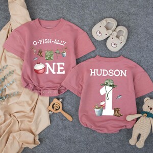 May include: Two dusty rose short-sleeved baby rompers. One features a fishing graphic with the text O-FISH-ALLY ONE. The other features a large number 1, a fishing hat, and the name HUDSON. Includes a pair of beige baby shoes and a wooden rattle.