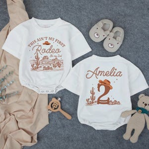May include: Two white baby rompers with a western theme. One says "This ain't my first Rodeo" with a desert scene. The other says "Amelia" and the number "2". Also pictured are bear slippers, a wooden rattle, and a stuffed bear.