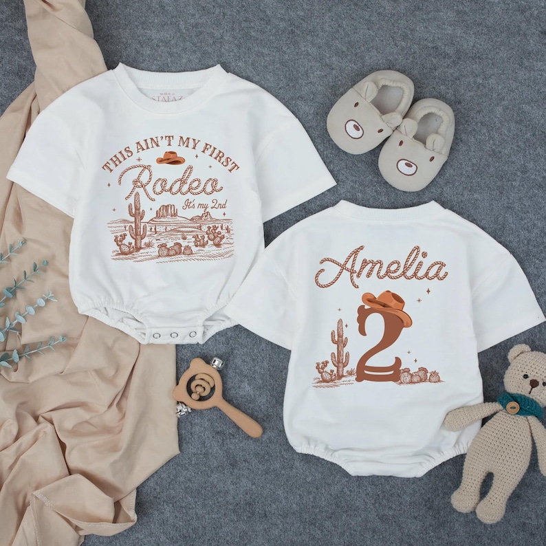 May include: Two white baby rompers with a western theme. One says "This ain't my first Rodeo" with a desert scene. The other says "Amelia" and the number "2". Also pictured are bear slippers, a wooden rattle, and a stuffed bear.