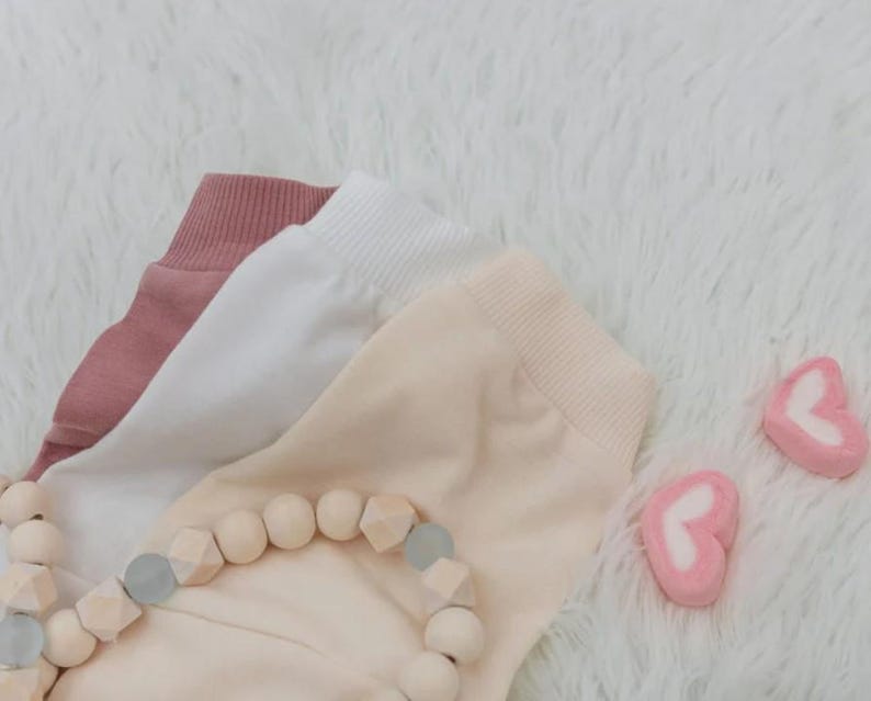Puede incluir: Tres pares de pantalones de beb&eacute; en rosa, blanco y melocot&oacute;n, con pu&ntilde;os acanalados. Un collar de cuentas de madera con cuentas geom&eacute;tricas y dos malvaviscos en forma de coraz&oacute;n tambi&eacute;n est&aacute;n en la imagen.