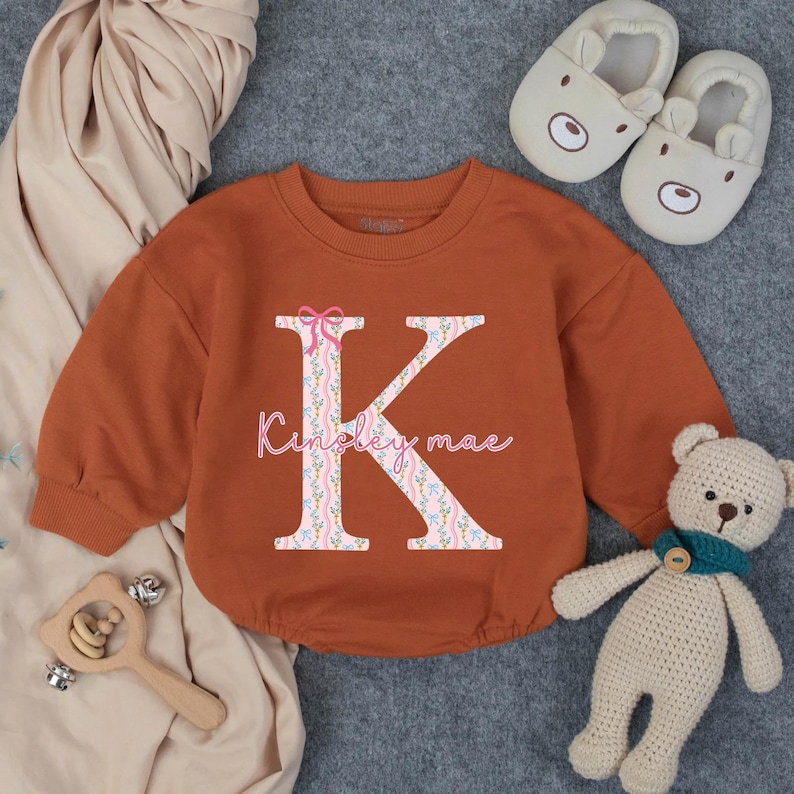 May include: Rust-colored baby romper with a large floral "K" initial and the name "Kinsley mae" in pink script. The romper is surrounded by a beige blanket, bear-shaped slippers, a wooden rattle, and a small teddy bear.