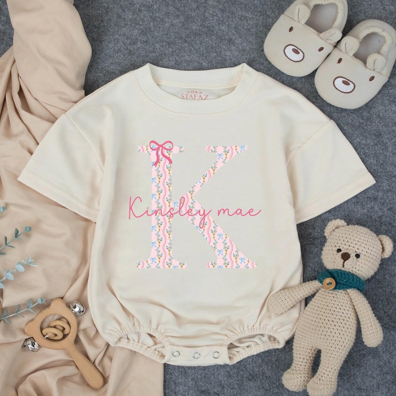 May include: Cream-colored baby romper with a large floral letter "K" and the name "Kinsley mae" in pink script. The romper has short sleeves and snap closures. A wooden rattle, teddy bear, and bear-shaped slippers are also in the image.