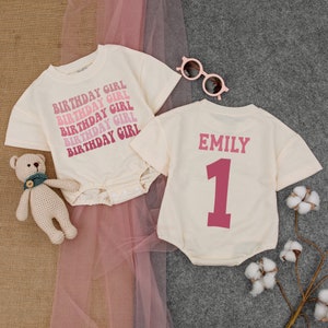 May include: Two white baby onesies. One onesie has the text "Birthday Girl" repeated in pink and red. The other onesie has the name "Emily" and the number "1" in pink.