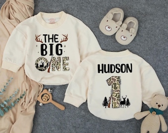 May include: Two cream-coloured baby sweatshirts with long sleeves. One says "THE BIG ONE" with a deer and camouflage design. The other says "HUDSON" and "1" with a deer, trees, and camouflage design. Also pictured are bear slippers and a teddy bear.