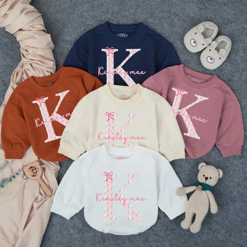 May include: Five long-sleeved sweatshirts in various colors: navy, rust, cream, mauve, and white. Each sweatshirt features a large letter "K" with a floral design and the name "Kinsley mae" in pink script. A teddy bear and baby slippers are also in the image.