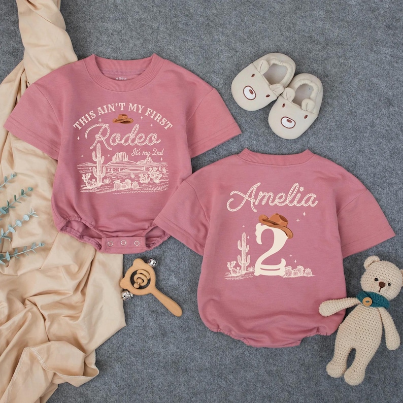 May include: Two pink baby bodysuits with short sleeves. One says "This ain't my first Rodeo" with a desert scene graphic. The other says "Amelia 2" with a cowboy hat and cactus. Includes bear slippers, a wooden rattle, and a teddy bear.