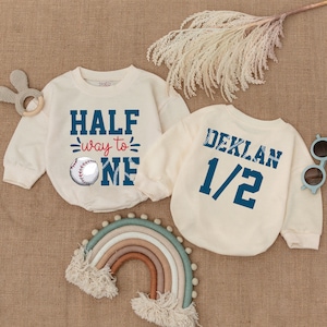 May include: Two cream-colored long-sleeved shirts with baseball-themed graphics. One shirt says "Half way to one" with a baseball graphic. The other shirt says "Deklan 1/2".