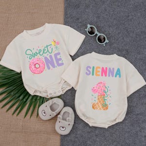 May include: Two cream-colored baby onesies with short sleeves. One says "Sweet ONE" with a donut graphic. The other says "SIENNA" and features a number one ice cream cone graphic. Gray baby shoes and sunglasses are also in the image.