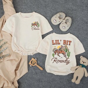 May include: Two cream-colored baby rompers with short sleeves, featuring a cowboy-themed graphic. One says "Olivia" and the other says "Lil' Bit Rowdy." The image also includes bear-shaped slippers, a wooden rattle, and a small teddy bear.