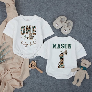 Puede incluir: Body de beb&eacute; blanco con el texto "ONE lucky duck!" y un gr&aacute;fico de pato. El n&uacute;mero "1" en la parte posterior est&aacute; en estampado de camuflaje, con el nombre "MASON" encima. Incluye una sonaja de madera y un oso de peluche.