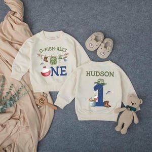 May include: Two cream-colored long-sleeve sweatshirts featuring fishing-themed graphics. One displays the text O-FISH-ALLY ONE with a fishing bobber, while the other shows the name HUDSON above a large blue number one and fishing gear.