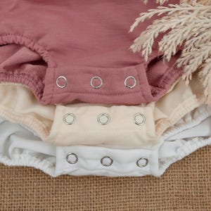 May include: Close-up of three baby bodysuits in dusty rose, cream, and white. Each bodysuit has a snap closure at the bottom. The fabric appears soft and comfortable, suitable for infants. The background is a neutral burlap.