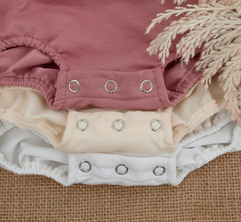 May include: Close-up of three baby bodysuits in dusty rose, cream, and white. Each bodysuit has a snap closure at the bottom. The fabric appears soft and comfortable, suitable for infants. The background is a neutral burlap.