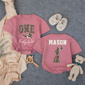 Puede incluir: Dos mamelucos rosas para beb&eacute; con mangas cortas. Uno tiene "ONE lucky duck!" y un gr&aacute;fico de pato. El otro tiene "MASON" y un gr&aacute;fico de pato con el n&uacute;mero "1". Tambi&eacute;n se muestran zapatos de beb&eacute; con forma de oso, una sonaja de madera y un oso de peluche.