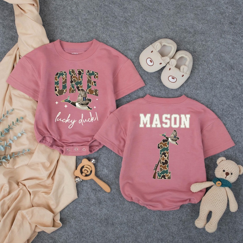Puede incluir: Dos mamelucos rosas para beb&eacute; con mangas cortas. Uno tiene "ONE lucky duck!" y un gr&aacute;fico de pato. El otro tiene "MASON" y un gr&aacute;fico de pato con el n&uacute;mero "1". Tambi&eacute;n se muestran zapatos de beb&eacute; con forma de oso, una sonaja de madera y un oso de peluche.
