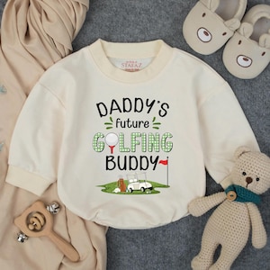 May include: Cream-colored baby onesie with long sleeves. The front features the text "DADDY'S future GOLFING BUDDY" in black and green. A golf-themed graphic includes a golf ball, golf cart, and flag.