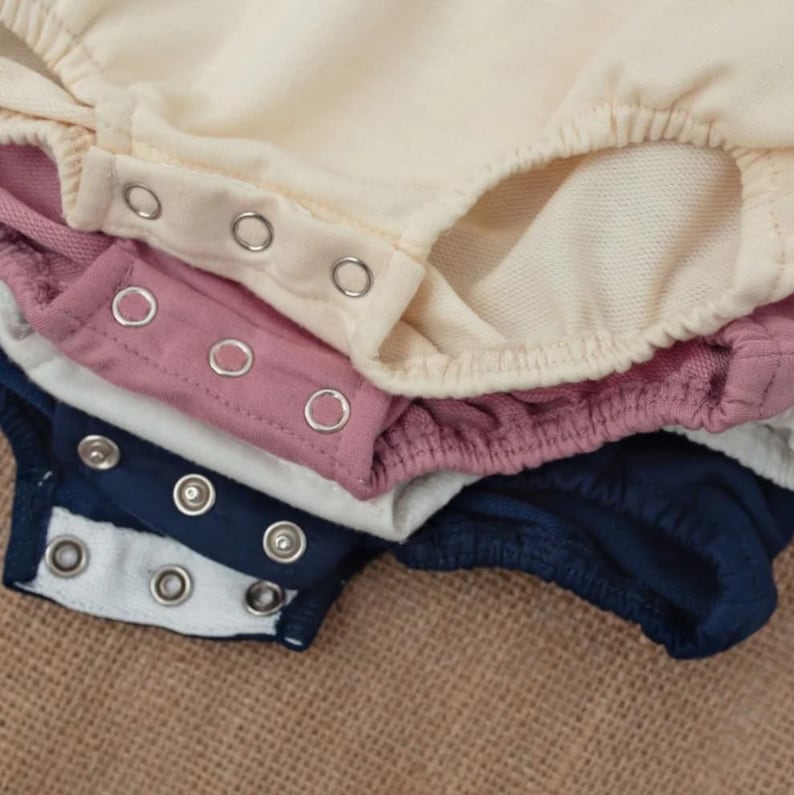 Puede incluir: Primer plano de varios bodies de beb&eacute; en crema, rosa empolvado, blanco y azul marino. Cada body tiene cierres de presi&oacute;n y aberturas el&aacute;sticas para las piernas. La tela parece suave y c&oacute;moda.