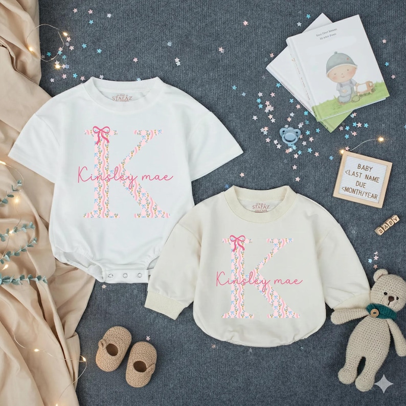 May include: Two baby bodysuits, one white and one cream, each with a floral "K" and the name "Kinsley mae". A teddy bear, baby shoes, and a book are also in the image. The text "BABY" is visible on a small sign.
