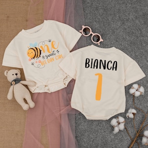 May include: Two white onesies with short sleeves. One onesie has a bee with the text "one sweet bee-day girl". The other onesie has the name "Bianca" and the number "1" in orange.