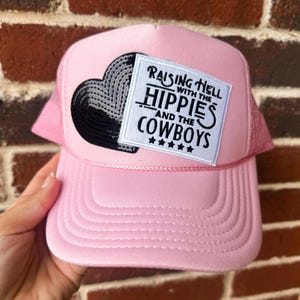 May include: Pink trucker hat with a mesh back and a curved brim. The hat features a patch with a black heart design and the text "Raising Hell with the Hippies and the Cowboys."