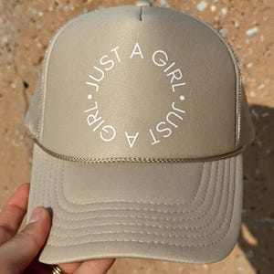 May include: A beige trucker hat with a white embroidered circle that says "Just a Girl. Just a Girl."