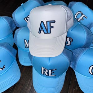 May include: A group of ten blue and white trucker hats with different initials embroidered on the front. The hats are made of a soft, breathable material and have a mesh back for ventilation.