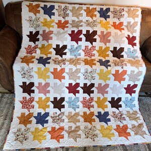 May include: A rectangular quilt featuring a repeating pattern of maple leaf shapes in various colors and fabric prints, including solid mustard yellow, dark brown, maroon, and light blue, alongside floral and polka dot patterns on a white background.