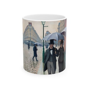 May include: A white ceramic mug with a painting of people walking in a Parisian street on a rainy day. The people are wearing coats and carrying umbrellas. The buildings in the background are tall and have many windows.