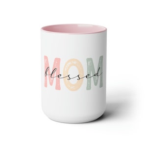 以下が含まれることがあります： ピンク色の内側が特徴の白いセラミックマグカップ。「MOM」の文字がパステルカラーでプリントされ、「blessed」の文字が筆記体で書かれています。シンプルでエレガントなデザインのギフトです。