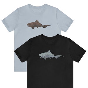 May include: Two t-shirts featuring different coloured prehistoric fish designs. The left shirt is light blue with a brown fish. The right shirt is black with a grey fish.
