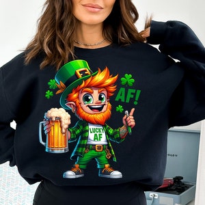 Könnte beinhalten: Schwarzes Sweatshirt mit einer Cartoon-Illustration eines Kobolds, der ein Bierglas hält. Der Kobold trägt einen grünen Hut, eine grüne Hose und ein kariertes Hemd. Der Text "Lucky AF!" ist auf dem Shirt aufgedruckt.