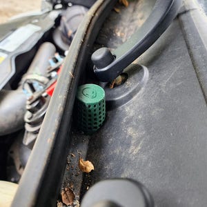 May include: A green, cylindrical automotive component featuring a grid design and a smiling face on its top surface. The part is situated near a black hinge and other car elements. The image is a close-up, emphasizing the component's details.