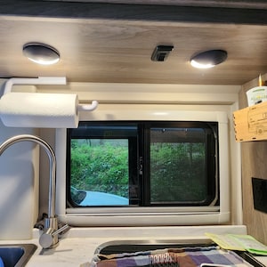 May include: Interior of a camper van kitchen, featuring a white paper towel holder with a roll of paper towels, a stainless steel faucet, and a window with a view of greenery. Two recessed lights are mounted overhead.