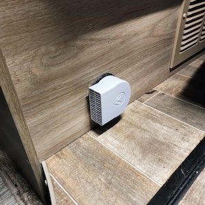 May include: A white, compact air vent with a circular design and a series of vertical slats. The vent is mounted on a wooden panel, near a floor with a wood-like pattern. The vent is likely part of a heating or cooling system.