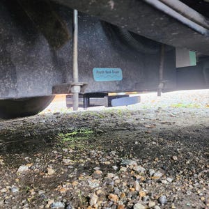 May include: Underside view of a recreational vehicle, showing the fresh water tank drain. A blue sticker with the words "Fresh Tank Drain" is visible. The image is taken from a low angle, with the ground and a tire in the foreground.