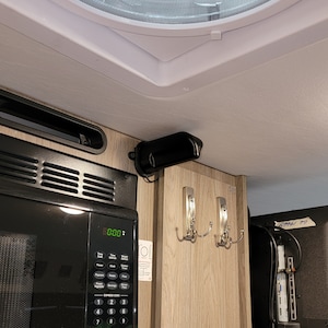 May include: Interior view of a recreational vehicle kitchen area. A black microwave oven is below a black vent hood. Two silver coat hooks are mounted on the light wood grain wall. A white ceiling vent is visible.